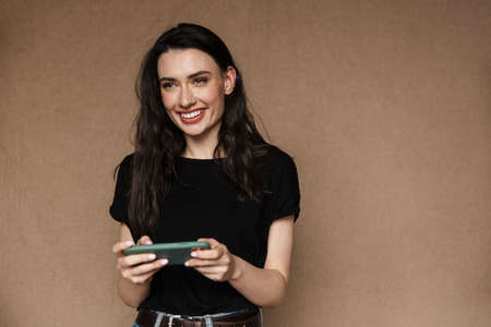 Photo of beautiful young brunette woman smiling and playing video games on cellphone isolated over...