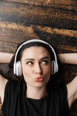 Photo from above of beautiful young brunette woman wearing headphones looking upward and lying on wooden floorの写真素材