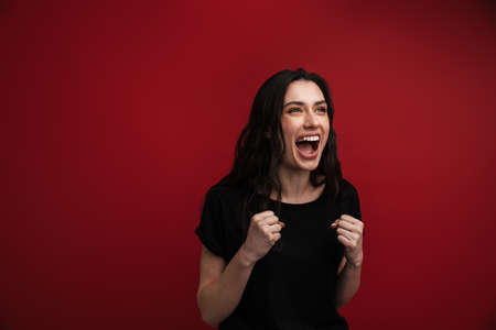 Photo of brunette excited woman in casual t-shirt winner gesture and screaming isolated over red backgroundの写真素材