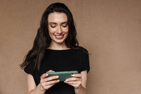 Photo of beautiful young brunette woman smiling and playing video games on cellphone isolated over...