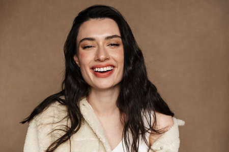 Photo of joyful young woman in jacket laughing and looking at camera isolated over beige backgroundの写真素材