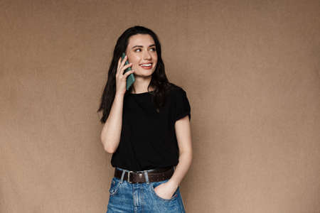 Photo of beautiful young brunette woman smiling and talking on cellphone isolated over beige...