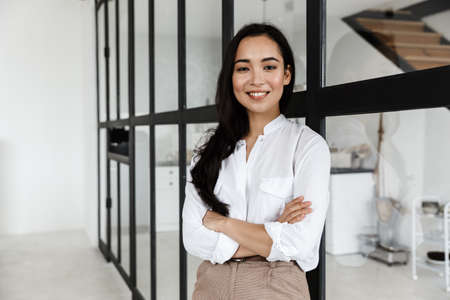 Beautiful smiling young asian woman wearing white shirt standing in a modern living roomの写真素材