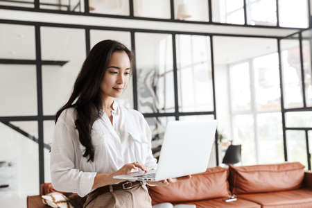 Attractive young brunette asian woman using laptop computer while standing in a stylish living room, wearing wireless earphonesの写真素材