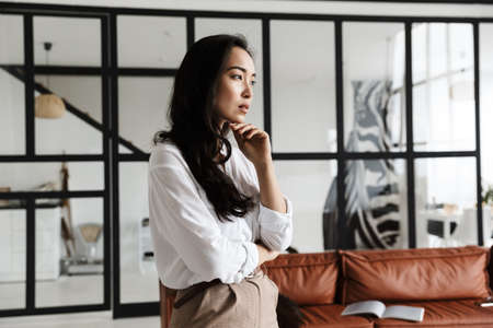 Beautiful young asian woman wearing white shirt standing in a modern living roomの写真素材