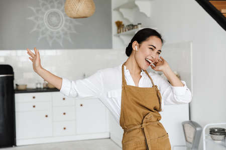 Smiling attractive young asian woman wearing apron dancing at the kitchen at home, listening to music with wireless earphonesの写真素材