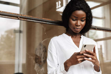 Attractive smart young african woman entrepreneur standing while leaning on a glass wall, using mobile phoneの写真素材