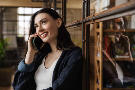 Attractive smiling smart young woman entrepreneur standing while leaning on a glass wall, talking on mobile phoneの写真素材