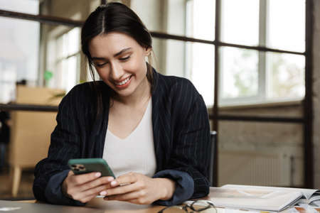 Confident smiling attractive young woman entrepreneur sitting at the office desk, using mobile phone