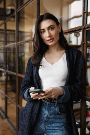 Attractive smart young woman entrepreneur standing while leaning on a glass wall, using mobile phone