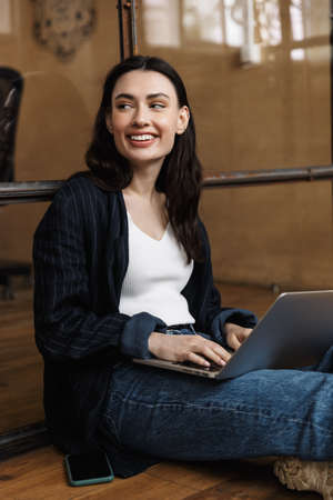 Attractive smiling young woman sitting at the office, leaning on a glass wall, using laptop computerの写真素材