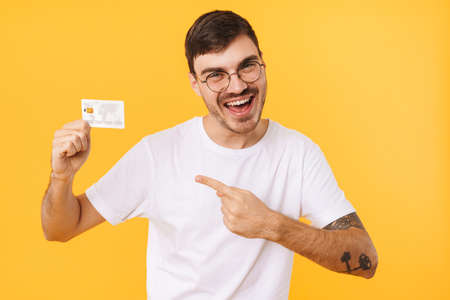 Photo of joyful young man in eyeglasses holding and pointing finger at credit card isolated over yellow backgroundの写真素材