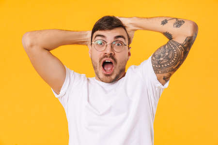 Photo of surprised young man in eyeglasses screaming and grabbing his head isolated over yellow backgroundの写真素材