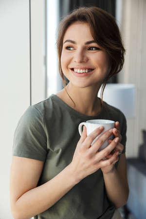 Attractive smiling young woman holding coffee cup while standing at the window indoorsの写真素材