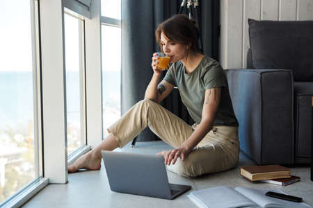 Attractive smiling young woman sitting at the couch on a floor of a living room, using laptop...
