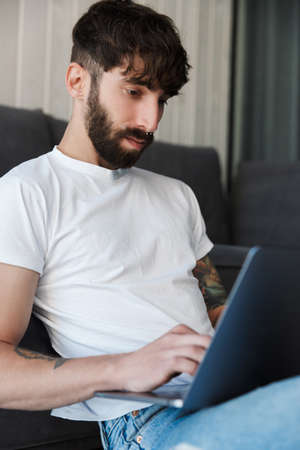 Handsome confident young bearded man using laptop computer while sitting at the couch on a floor in...