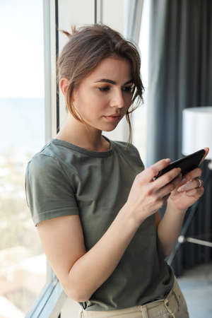 Attractive young woman using mobile phone while standing at the window indoors