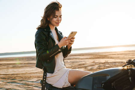 Beautiful young girl wearing leather jacket sitting on a motorbike at the sunny beach, using mobile...