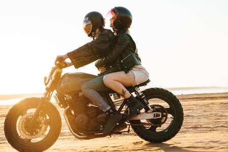 Side view of a beautiful young stylish couple enjoying ride on a motorbike at the beachの写真素材