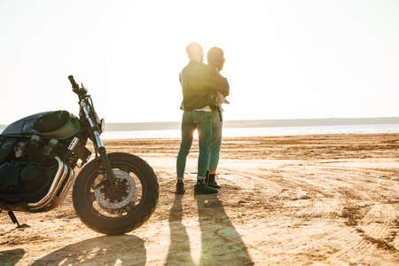 Beautiful young cople wearing jackets spending time at the sunny beach, standing near a motorbikeの写真素材