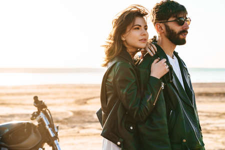 Attractive young couple embracing while standing near a motorbike at the sunny beachの写真素材
