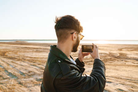 Back view of an stylish young bearded man taking picture on mobile phone while standing at the sunny beachの写真素材