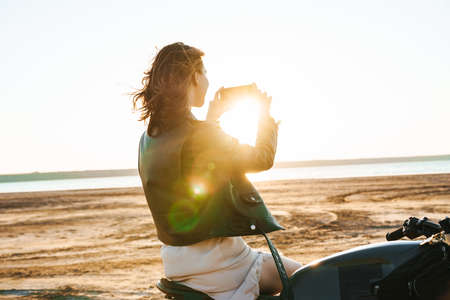 Beautiful young girl wearing leather jacket sitting on a motorbike at the sunny beach, taking a picture of a sunsetの写真素材