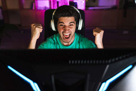 Image of excited young man in headphones making winner gesture while playing video game on computer at homeの写真素材