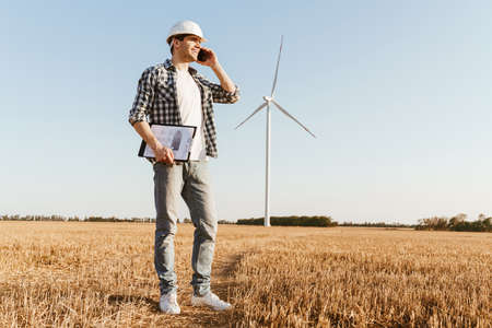 A smiling male engineer using mobile phone while standing at the electric windmill farmの写真素材