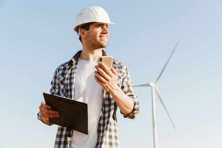 A smiling male engineer using mobile phone while standing at the electric windmill farm