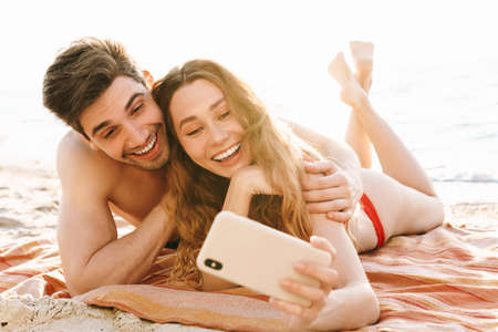 Happy young couple taking a selfie on the beach while laying on blanketの写真素材