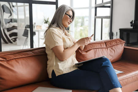 Image of mature business woman sitting on sofa indoors at home while using mobile phone.の写真素材