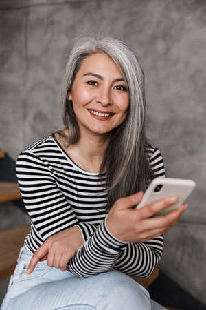 Image of cheerful positive mature woman sitting on steps indoors at home while using mobile phone.