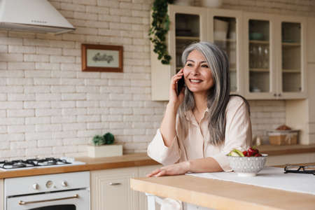 Picture of mature positive cheery woman sitting at the kitchen indoors at home talking by mobile phone.の写真素材