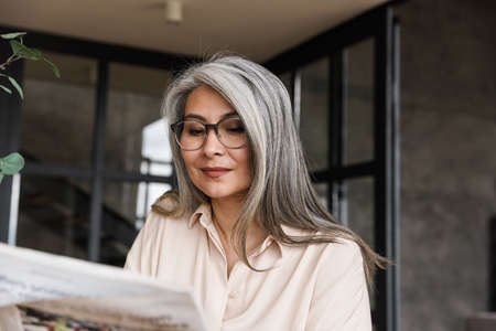 Image of mature concentrated woman indoors at home reading newspaper