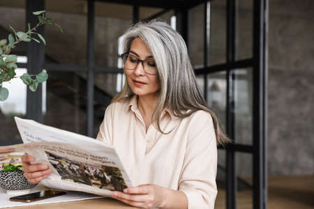 Photo of mature serious concentrated woman indoors at home reading newspaper