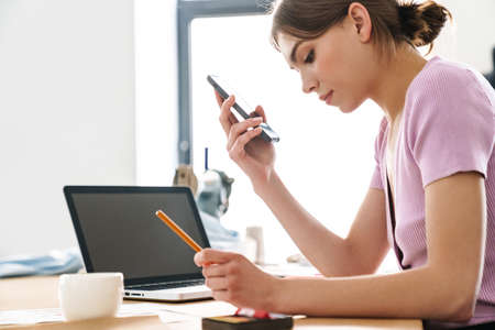 Photo of young focused woman using cellphone while working with laptop at table in office
