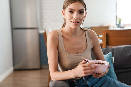 Attractive young woman having cereal breakfast on couch, smiling at camera