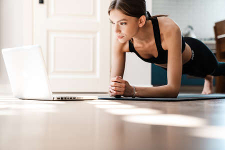 Attractive young sporty woman working out on a fitness mat at home, using laptop computerの写真素材