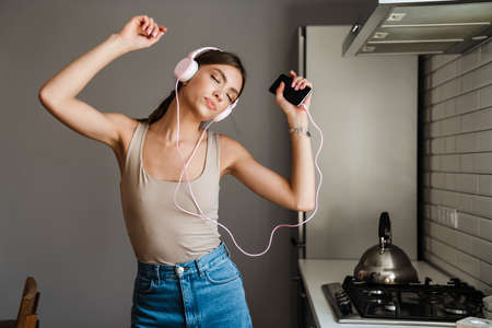 Young amazing emotional woman dancing in kitchen at home, listening to music with headphones, holding blank screen mobile phoneの写真素材
