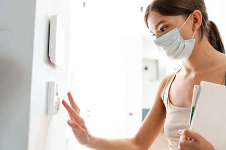 Young woman in medical mask entering code on keypad of home security alarm