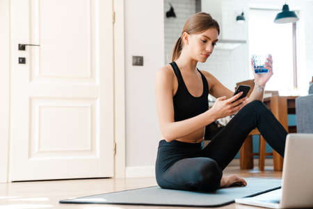 Attractive tired young fitness woman drinking water after work out while sitting on a mat with...