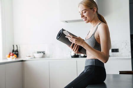 Photo of joyful beautiful woman with yoga mat using mobile phone while sitting on table in kitchen