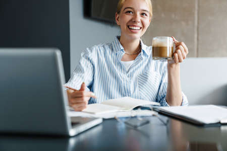 Photo of happy young woman drinking coffee and smiling while working with laptop at table in roomの写真素材