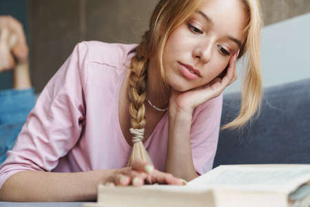 Photo of focused nice woman with blonde hair reading book while lying on sofa in bright roomの写真素材