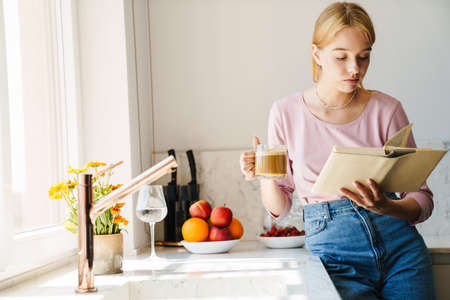 Photo of focused caucasian woman reading book while drinking coffee in modern kitchenの写真素材