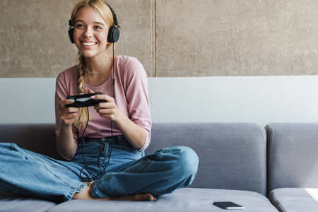 Photo of cheerful nice woman playing online game with joystick while sitting on couch in bright room