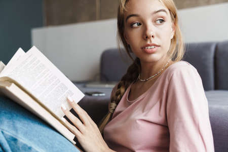 Photo of focused nice woman with blonde hair reading book while sitting on floor in bright room