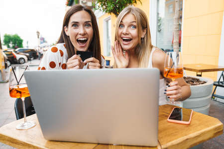 Image of delighted adult two women holding credit card while using laptop at table in cafe outdoorsの写真素材