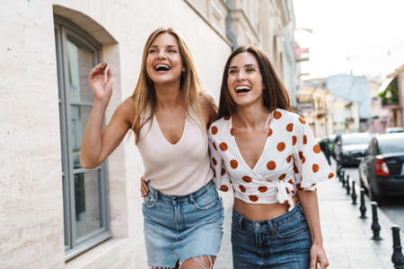 Image of cheerful adult two women hugging and laughing while walking on city street